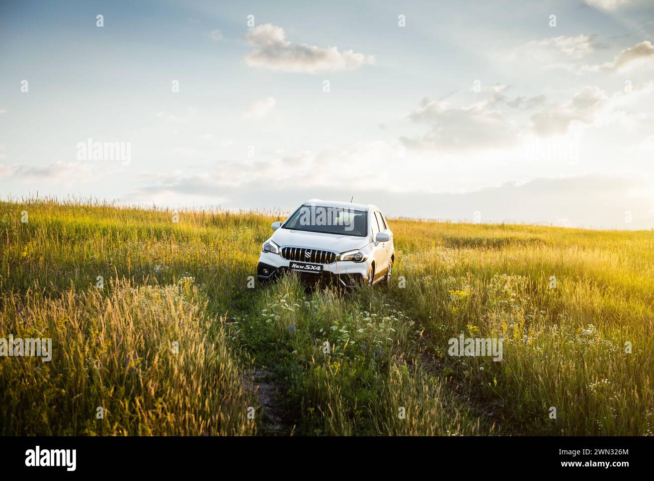 white Suzuki SX4 in hilly field at sunset. Front view of compact crossover in a green meadow - weekend away from the city Stock Photo