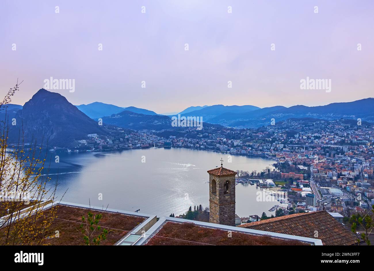 The mountain village of Aldesago opens the view on the mirror-like Lake Lugano and Monte San Salvatore against the purple twilight sky, Lugano, Ticino Stock Photo