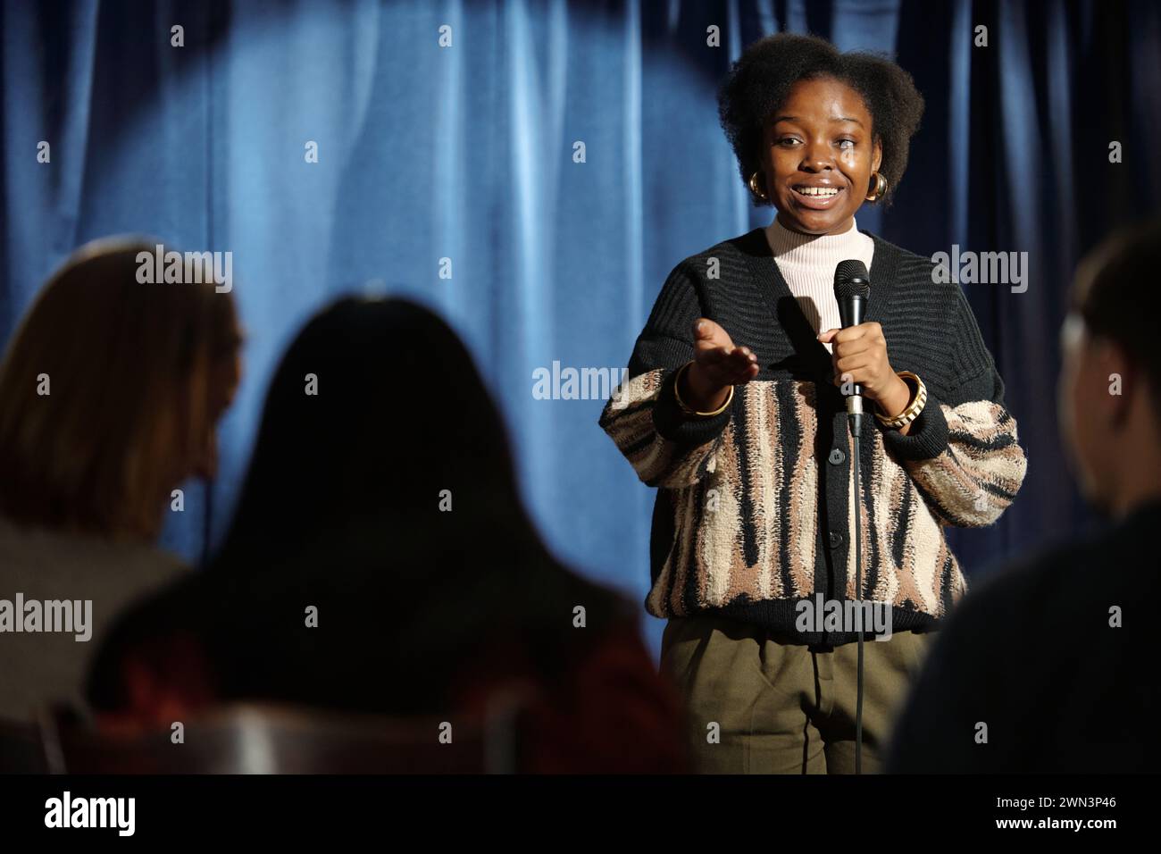 Young African American female comedian standing in front of audience on stage and speaking in