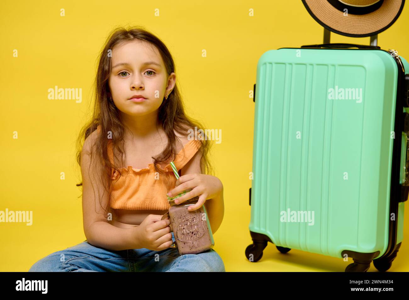 Lovely kid girl drinks a cocktail, sitting near a trendy cyan suitcase, looking confidently at the camera, isolated over yellow studio background. Peo Stock Photo