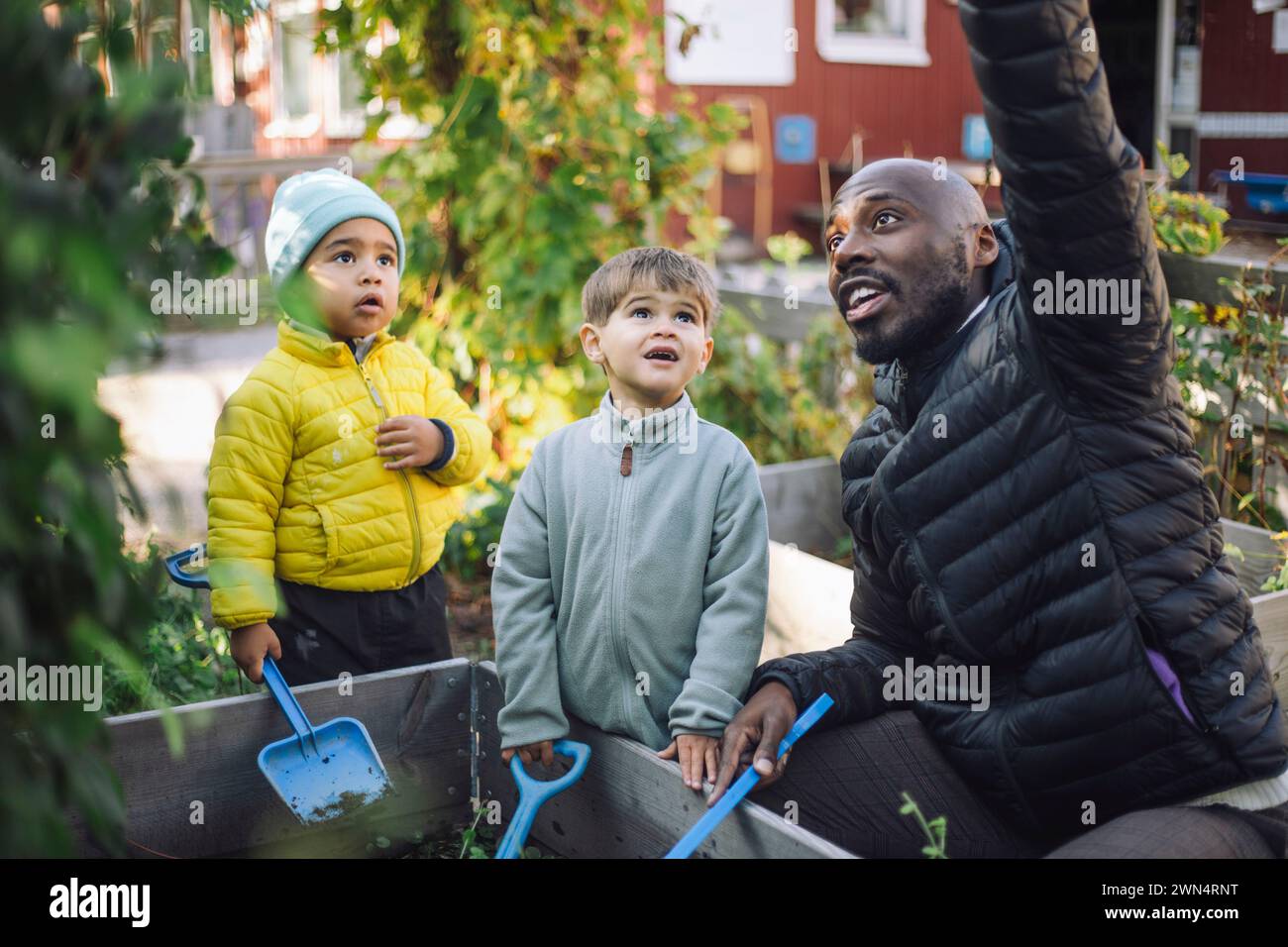 Male teacher talking with preschool kids holding shovel at garden Stock Photo