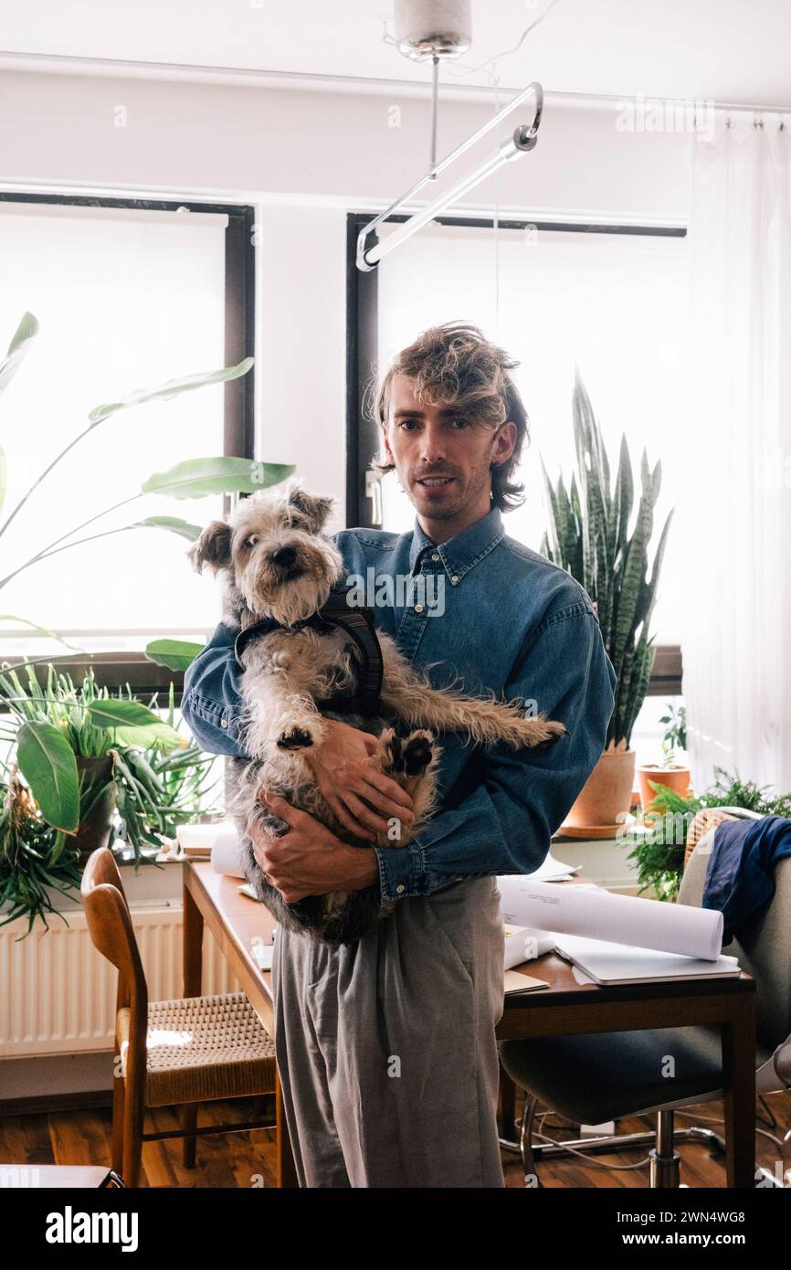 Portrait of male architect carrying dog standing at home office Stock Photo