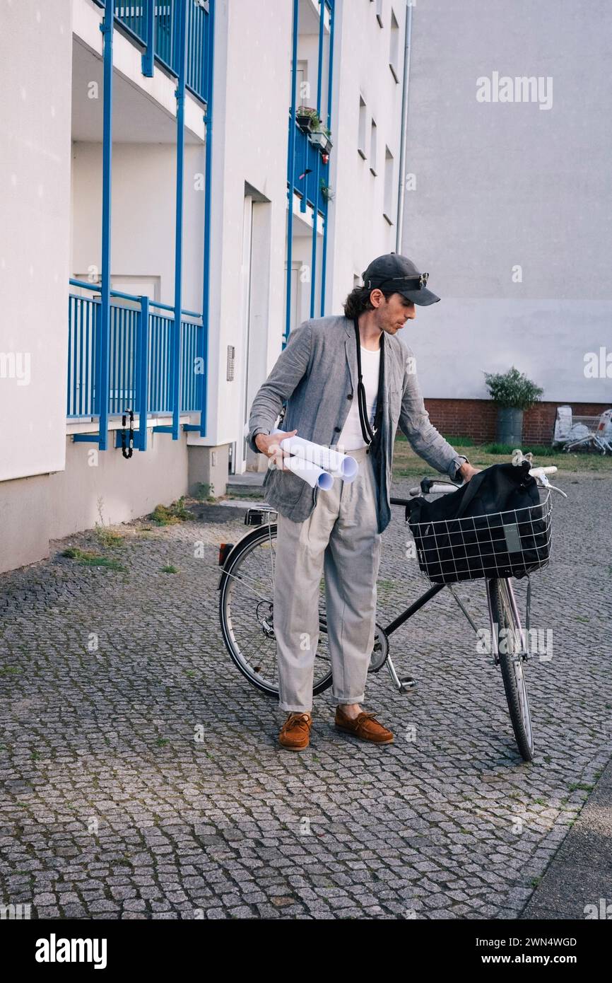 Full length of male architect holding blueprint rolls standing by bicycle on footpath Stock Photo