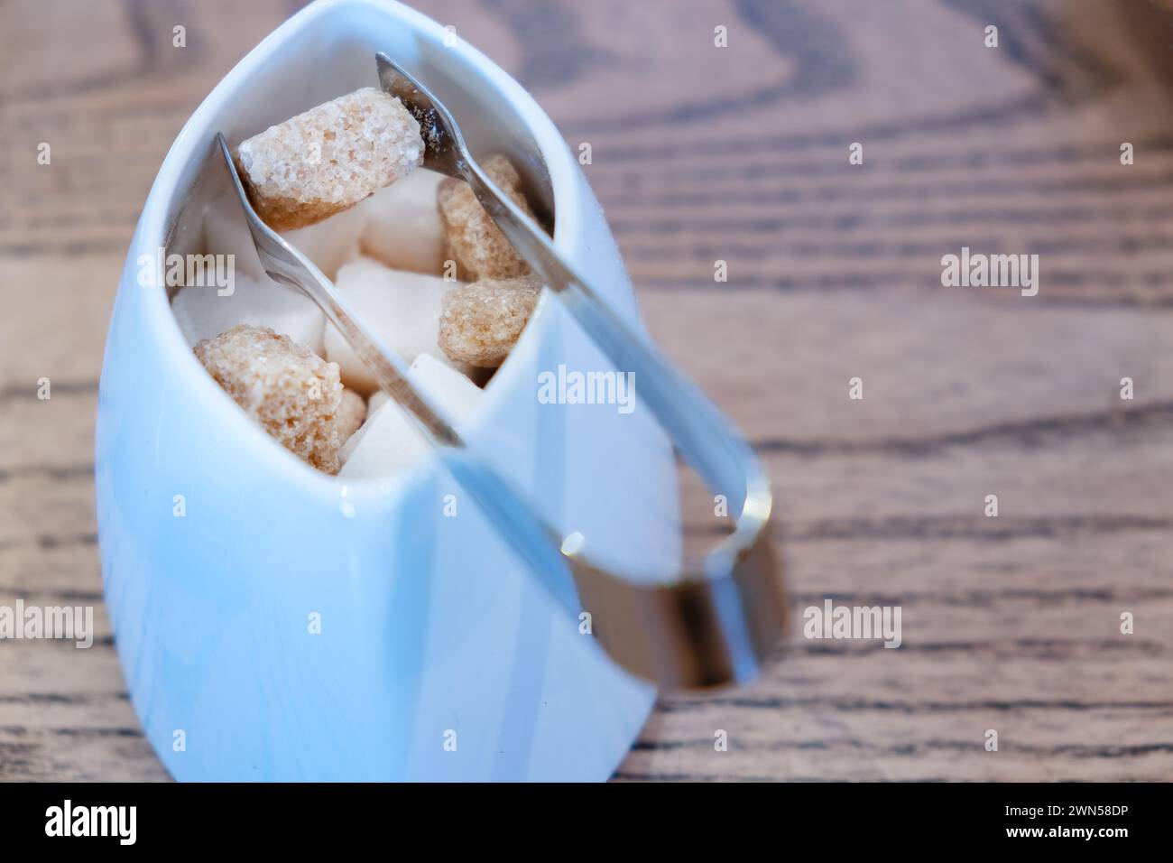 A small pair of sugar tongs gripping a brown sugar cube in a small sugar bowl.  The sugar is provided in a cafe to sweeten tea and coffee drinks Stock Photo