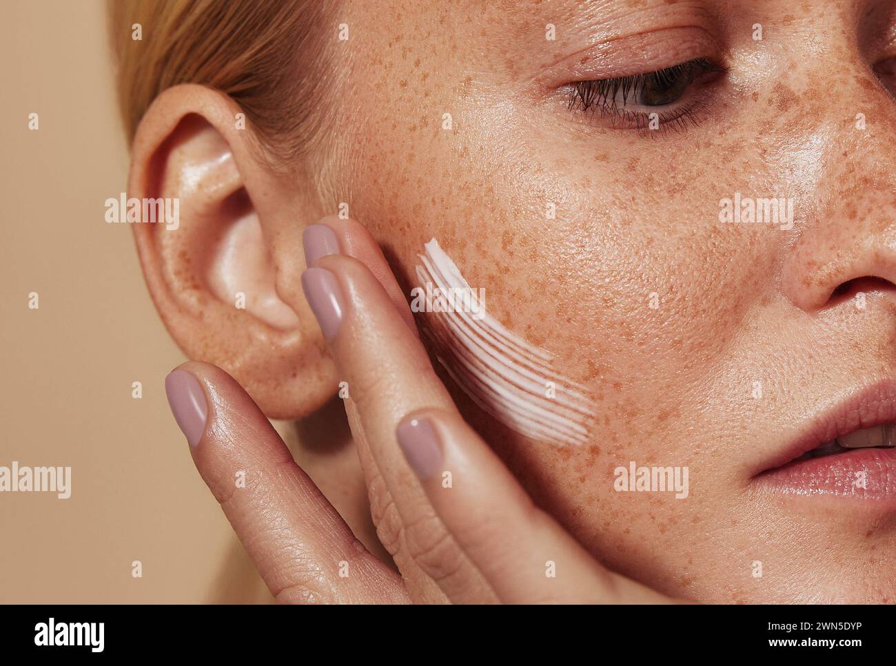Close up highly detailed shot of a young woman applying cream on her freckled skin. Cropped shot of woman applying moisturizer on cheek with fingers. Stock Photo