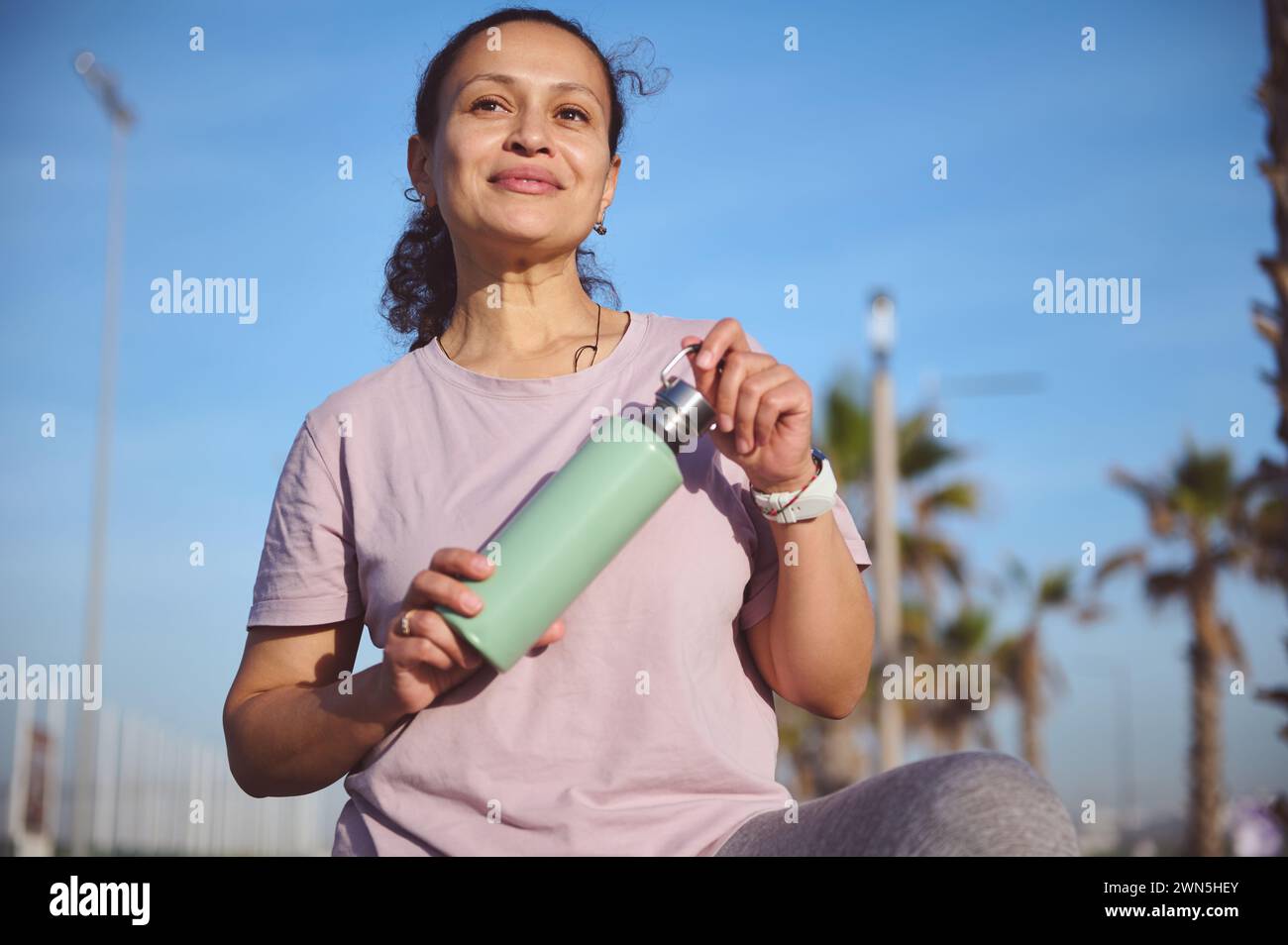 Attractive young sporty female, sportswoman, athlete holding a bottle with water, smiling, looking into distance, standing on the beach, resting after Stock Photo