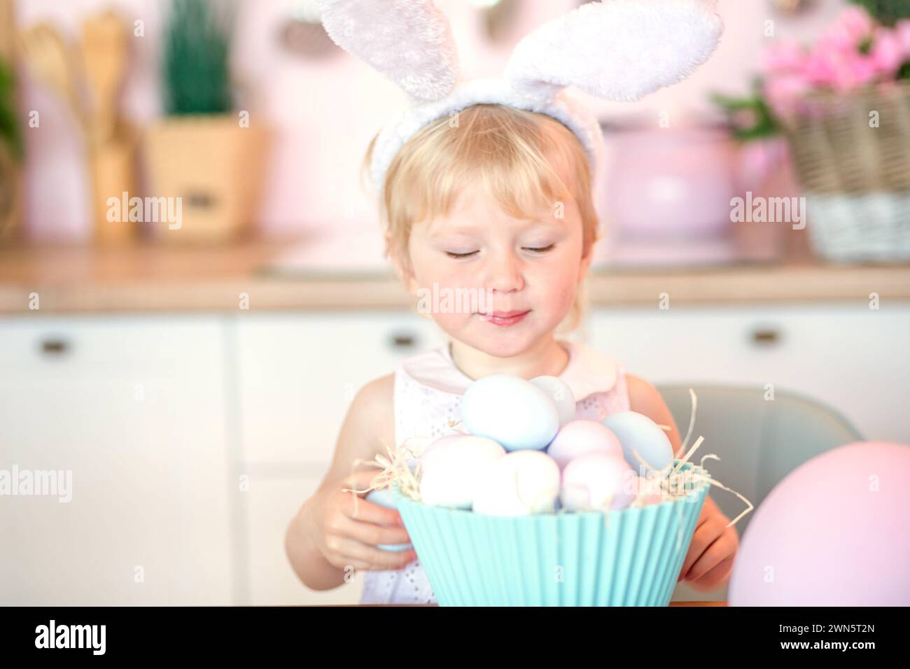 Little 3-year-old girl with plate full of painted easter eggs standing near table in kitchen decorated for Easter. Easter Egg Hunt Concept Stock Photo