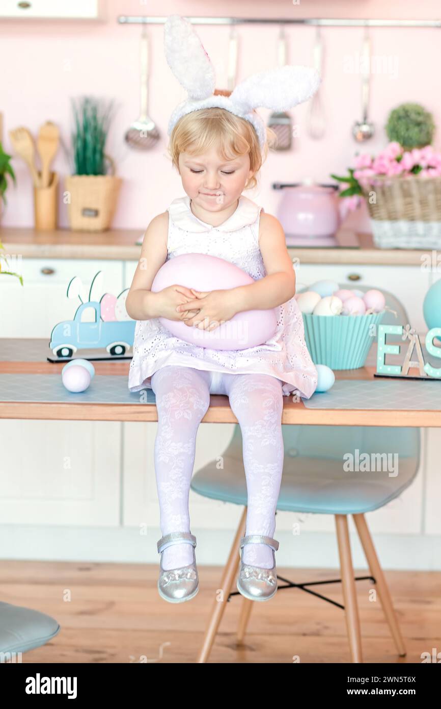 A little 3-year-old girl sits on a table in a kitchen decorated for Easter and holds a pink large egg in her hands. The concept of Easter egg hunt Stock Photo