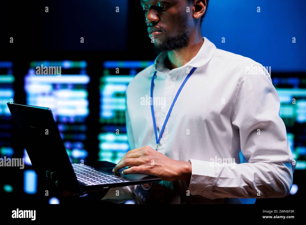 African american IT developer using laptop to examine server mainframes providing vast computing resources. Data center enabling AI to process massive datasets for complex machine learning operations Stock Photo