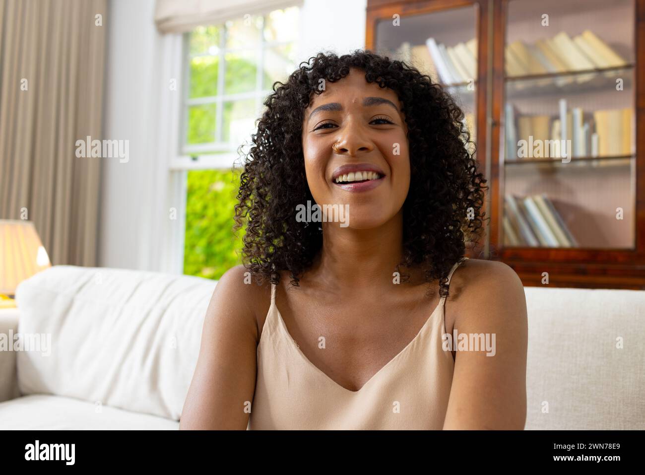 Young biracial woman with curly hair smiles warmly on a video call, seated on a white sofa Stock Photo