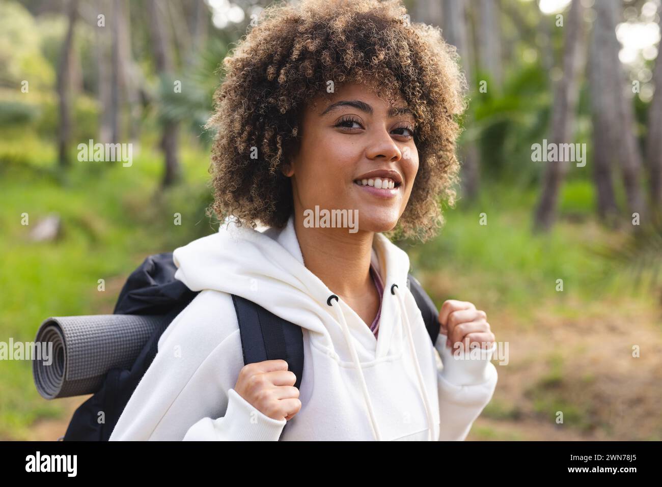 Young biracial woman with curly hair smiles while hiking in a forest on ...