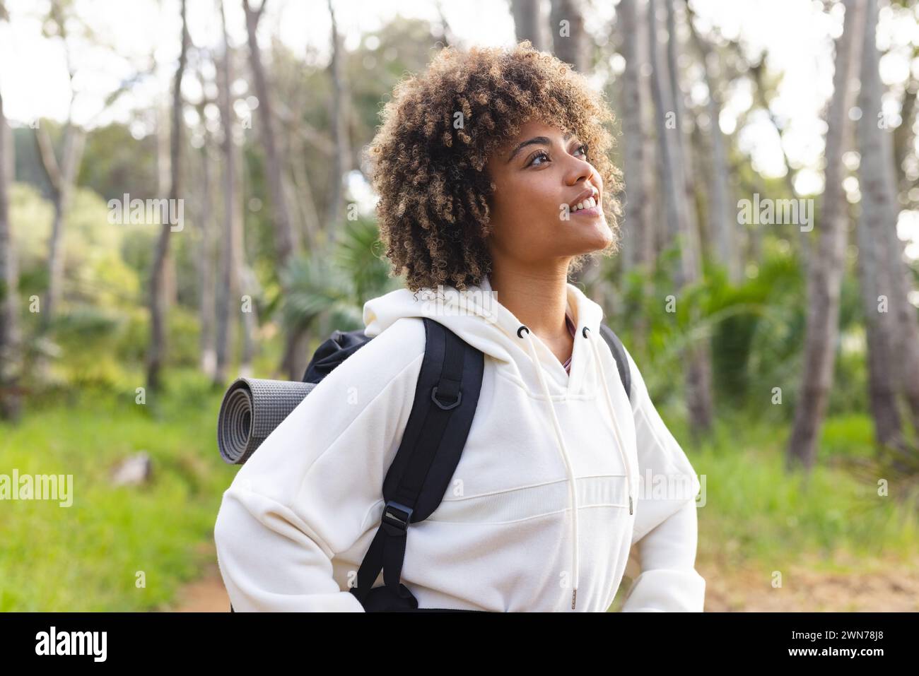 Young biracial woman with curly hair smiles while on a hike in a forest ...
