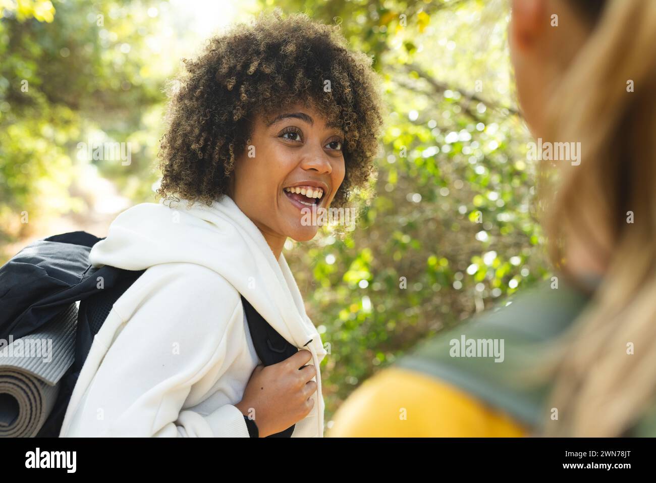 Young biracial woman with curly hair smiles brightly, carrying a backpack on a hike Stock Photo