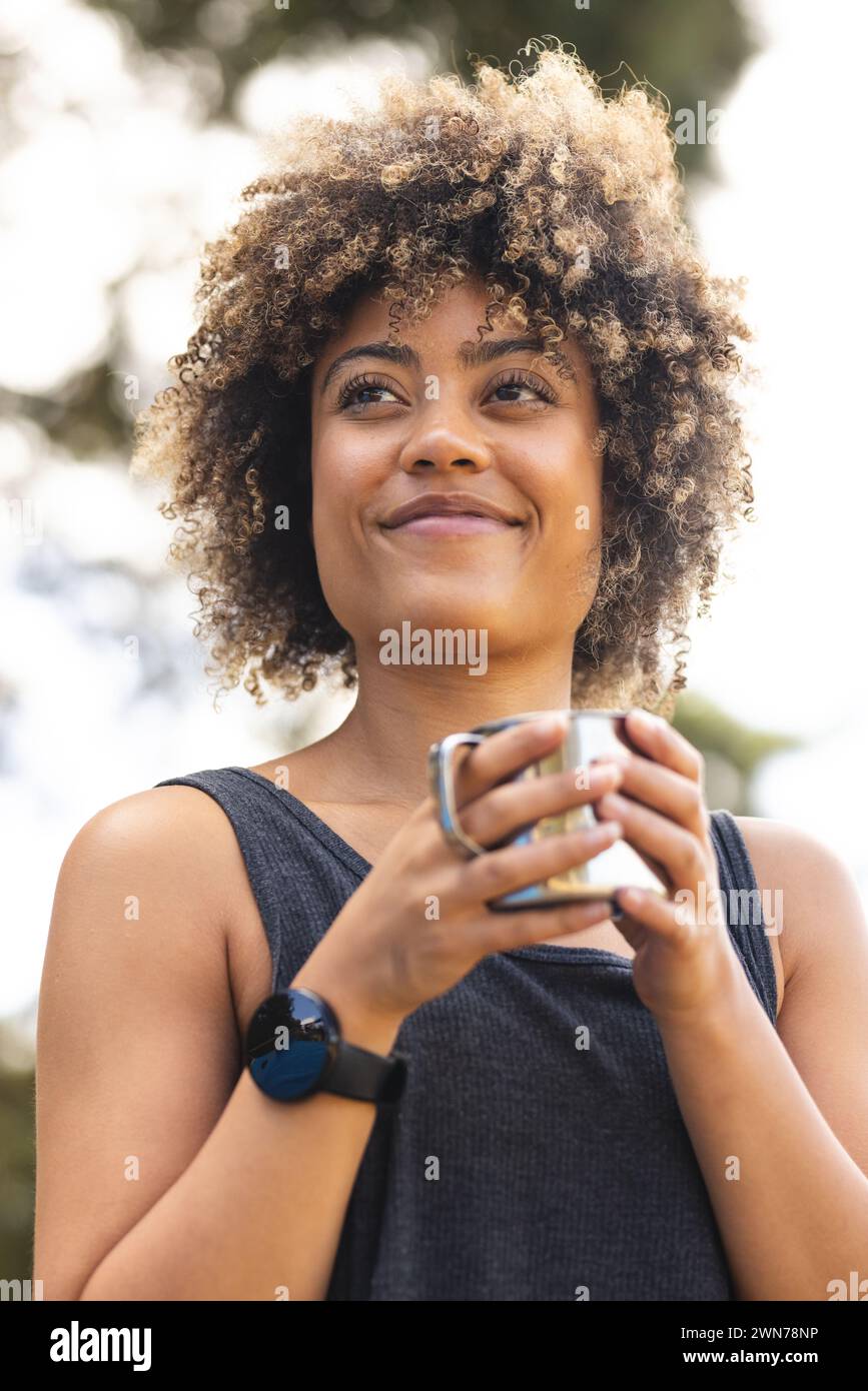 Young biracial woman with curly hair smiles while holding a cup outdoors Stock Photo