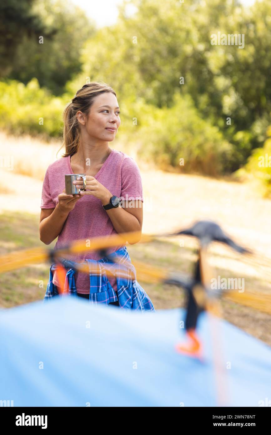 A young woman enjoys a cup of coffee near a tent in a sunny outdoor setting Stock Photo