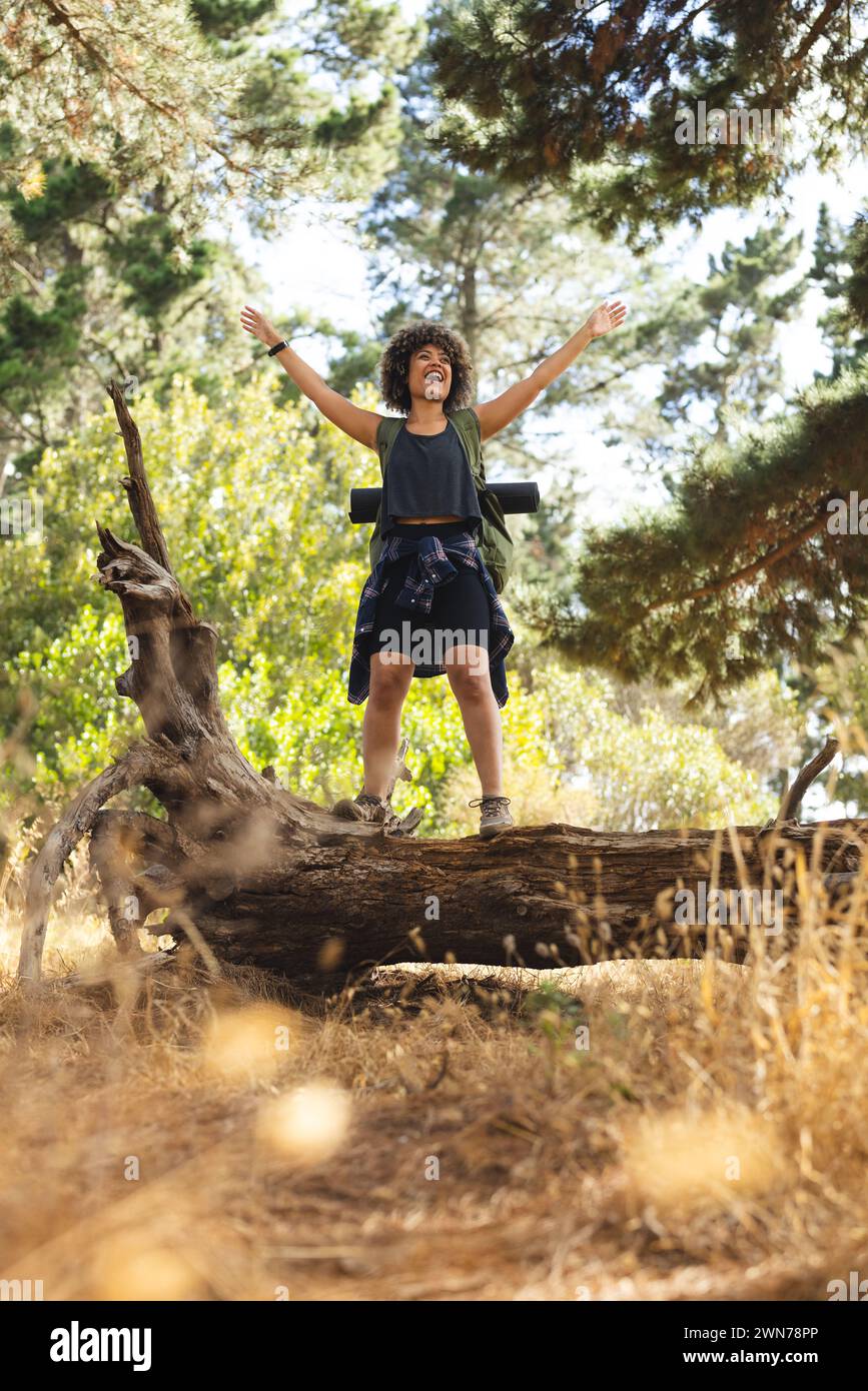 Young biracial woman stands on a fallen tree trunk, arms raised in joy, on a hike Stock Photo