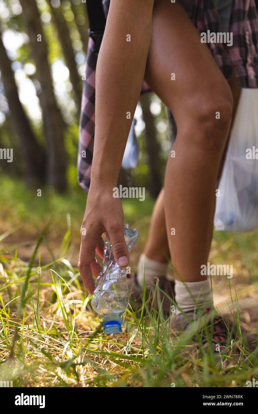 Person picks up a plastic bottle from the grass in a wooded area, collecting trash Stock Photo