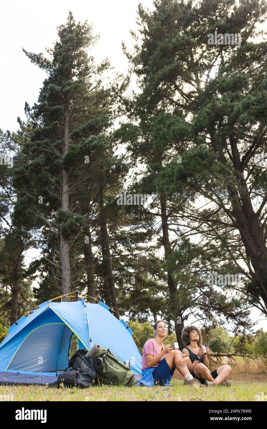 Two women relax beside a blue tent in a forest setting with copy space Stock Photo