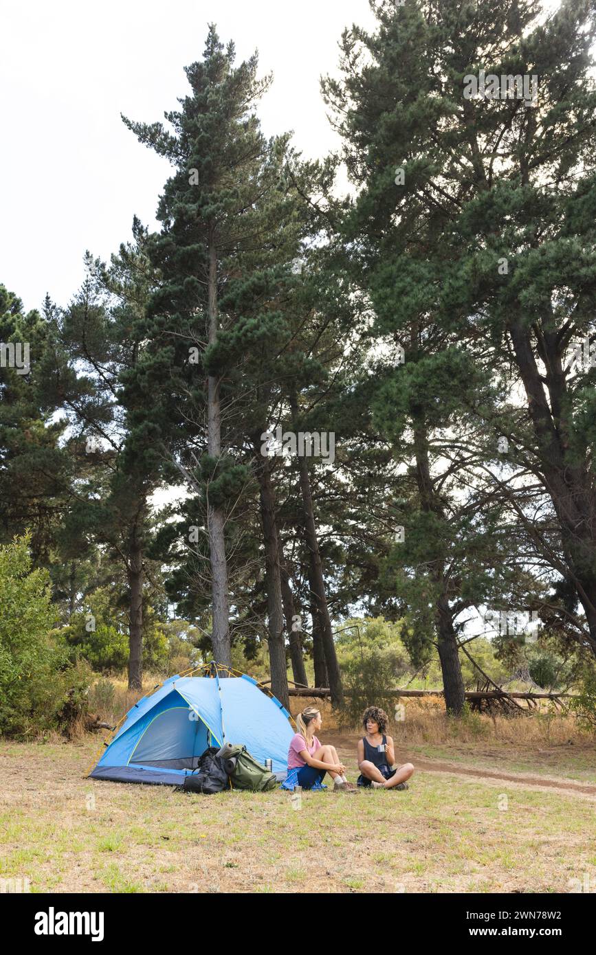 Two women relax beside a blue tent in a serene forest clearing with copy space Stock Photo
