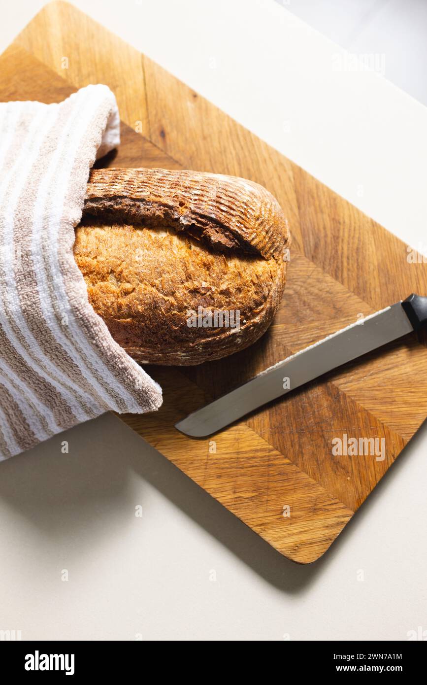 A loaf of bread and a knife rest on a wooden cutting board, partially covered by a striped towel Stock Photo