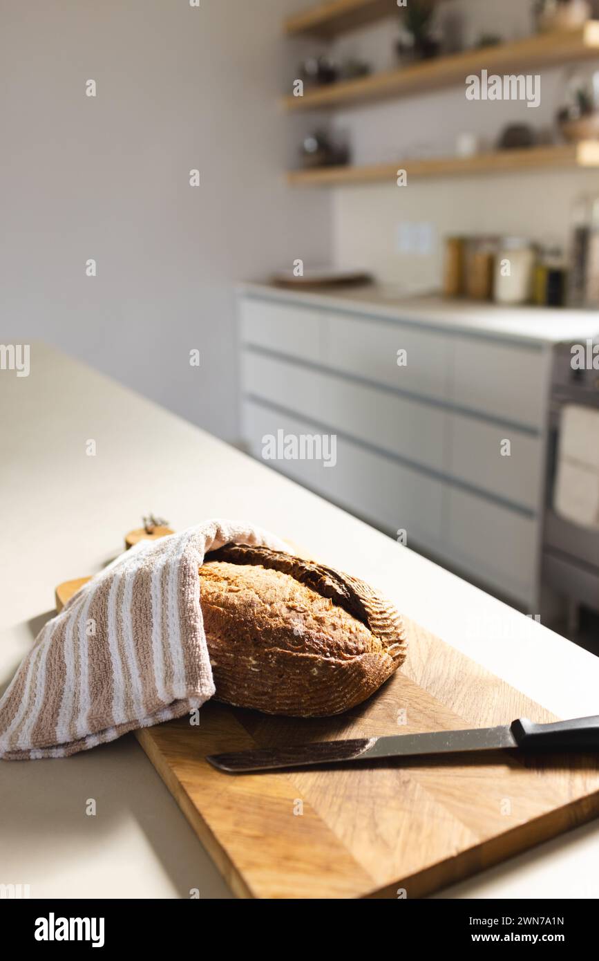 Sunlit kitchen scene with bread on board under towel, evoking warmth. Stock Photo