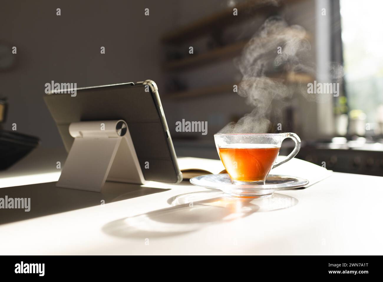 Steam rises from a hot cup of tea next to a tablet on a sunlit table Stock Photo