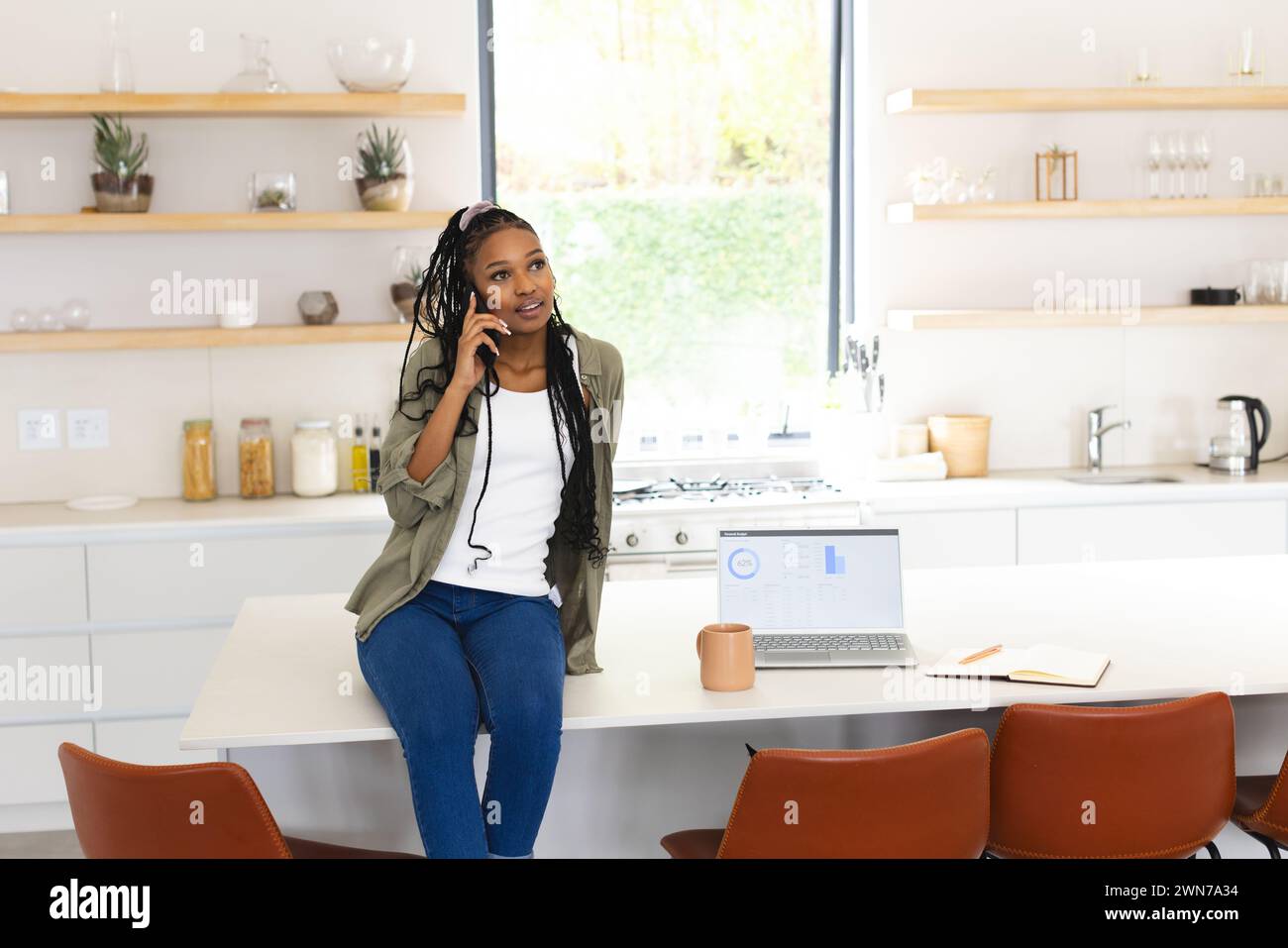 A young African American woman chats on the phone, leaning on a kitchen island with copy space Stock Photo