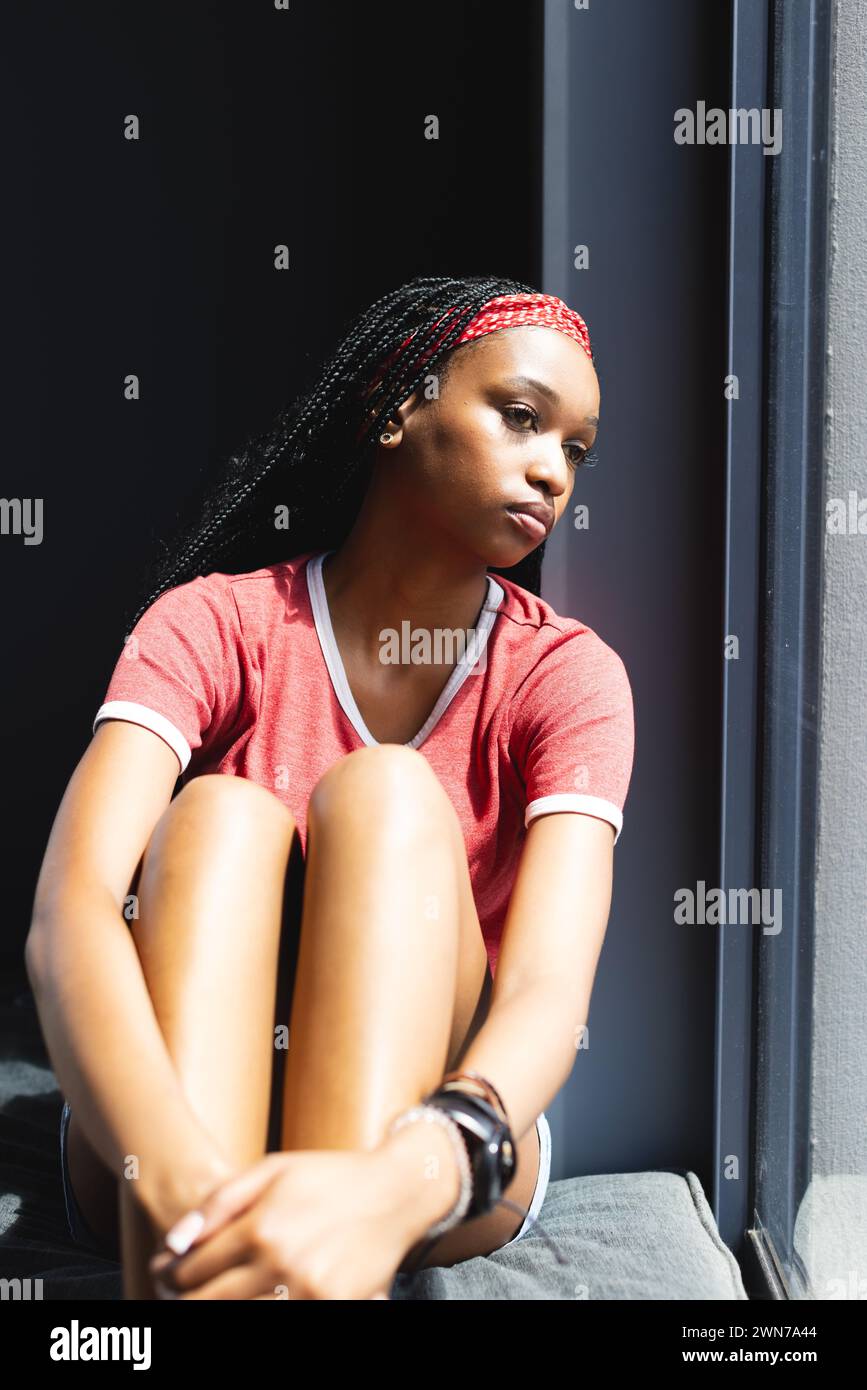 A young African American woman sits by a window, sunlight highlighting ...