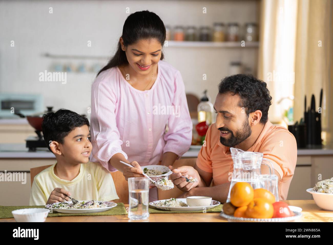 Happy Indian woman serving food to her son and husband on dining table at home - concept of family bonding, household responsibility and togetherness Stock Photo