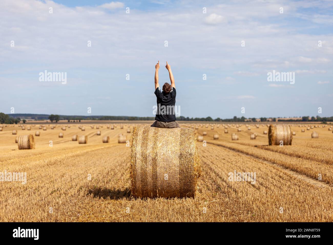 Ein Man sitz auf einem Strohballen und streckt die Arme in den Himmel vor Freude Stock Photo