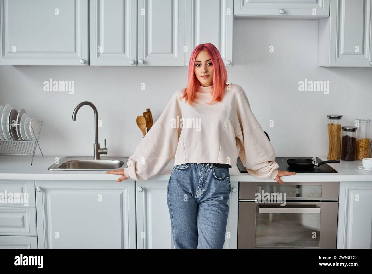 good looking jolly woman in casual jumper and jeans posing in kitchen and looking at camera Stock Photo