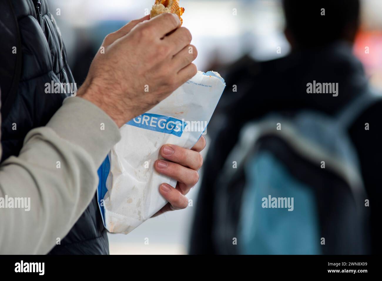 A Greggs bakery customer with a product in a carrier bag with the companies logo displayed as he enjoys the snack on a UK railway station.Greggs plc is a British bakery chain. It specialises in savoury products such as bakes, sausage rolls, sandwiches and sweet items including doughnuts and vanilla slices. It is headquartered in Newcastle upon Tyne, England. It is listed on the London Stock Exchange, and is a constituent of the FTSE 250 Index. Originally a high street chain, it has since entered the convenience and drive-thru markets. Stock Photo