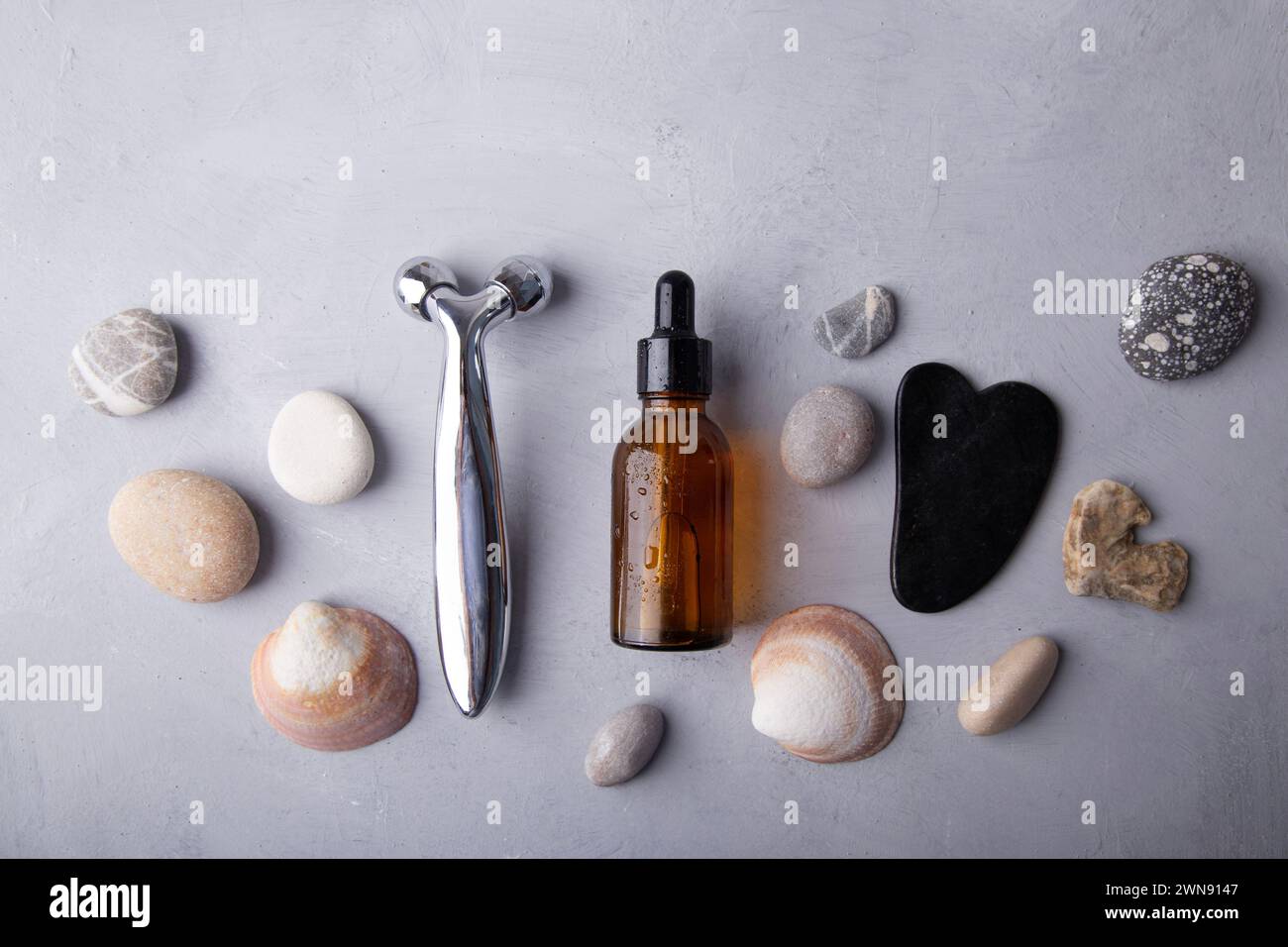 beauty essentials and facial massage tools among pebbles on a grey backdrop. Summer spa care concept. Stock Photo