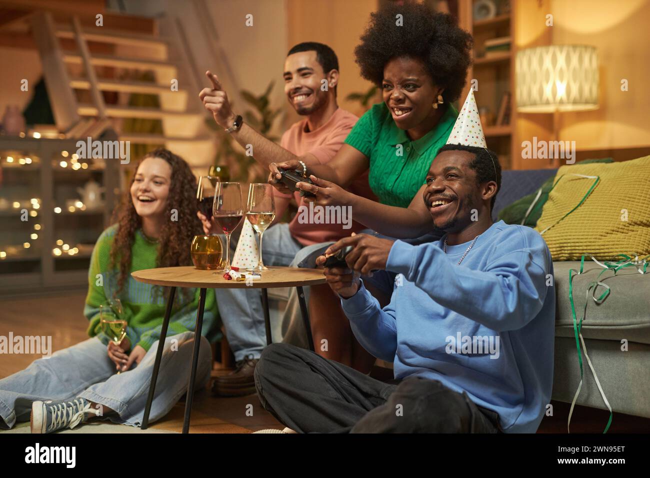 Full length portrait of multiethnic group of adults playing video games emotionally during party at home with African American woman holding controller Stock Photo