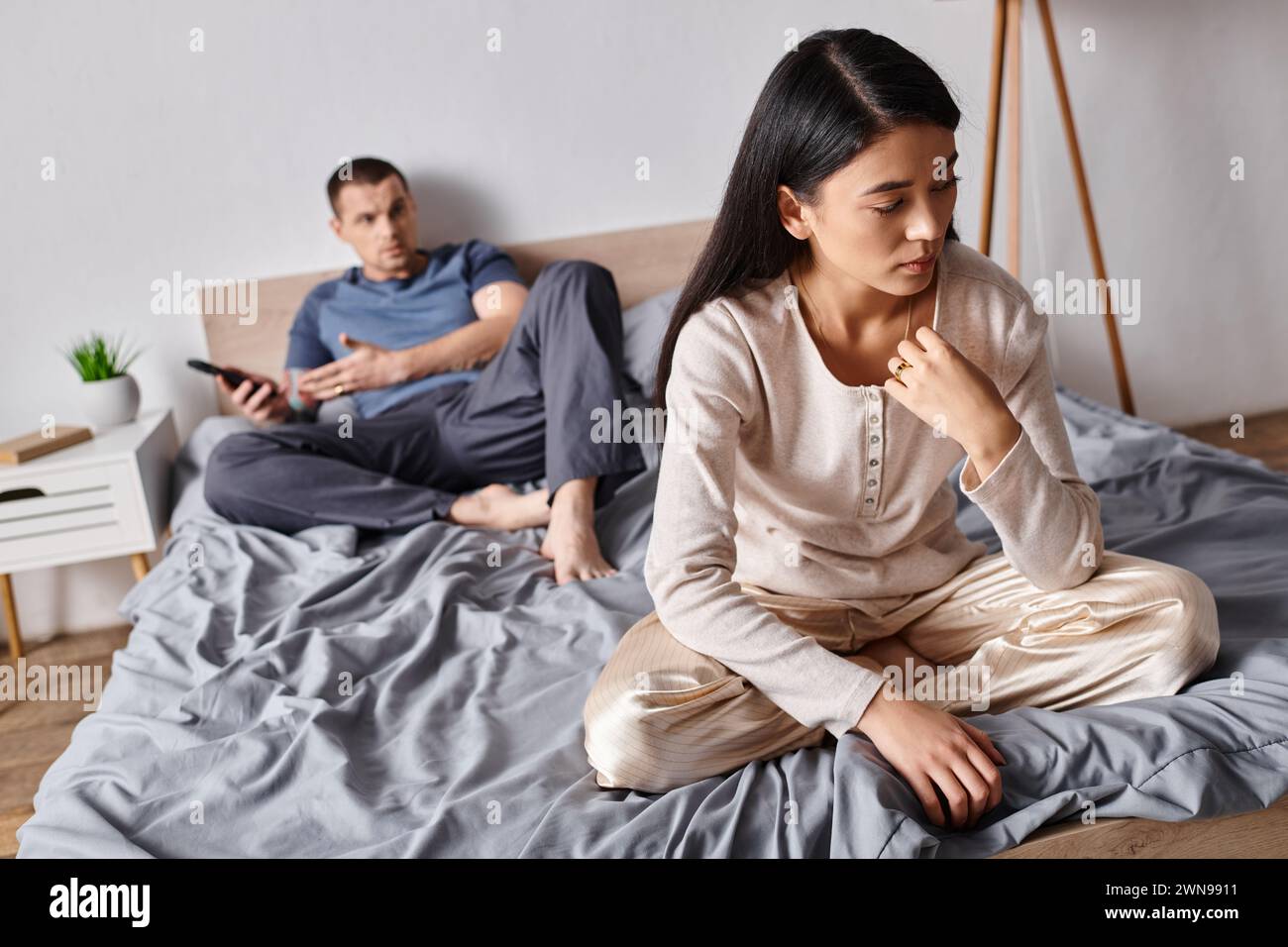 frustrated asian woman sitting near husband using smartphone in bedroom at home, family problem Stock Photo