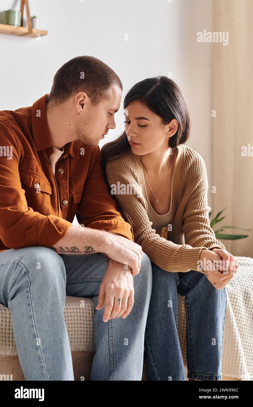 young and upset multiethnic couple sitting and talking on bed at home, relationship difficulties Stock Photo