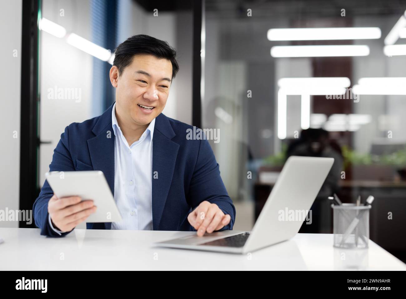 Modern businessman in formal clothes using digital tablet while working by pc on white table. Successful entrepreneur using two wireless devices for comparing articles from different web sources. Stock Photo