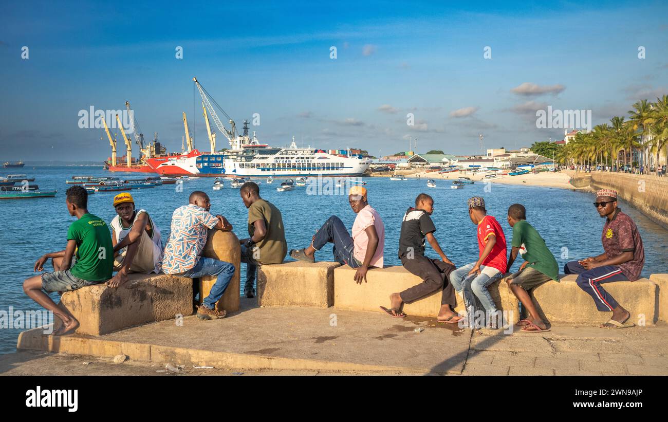 A group of men and boys sit on the sea wall near the ferry port in Stone Town, Zanzibar, Tanzania Stock Photo