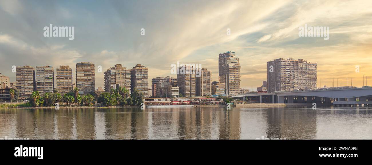 As the day comes to a close, the golden sun rays illuminate the Maadi skyline by the Nile. The calm waters reflect the city's silhouette, with Mounib Bridge visible in the distance, Cairo, Egypt Stock Photo