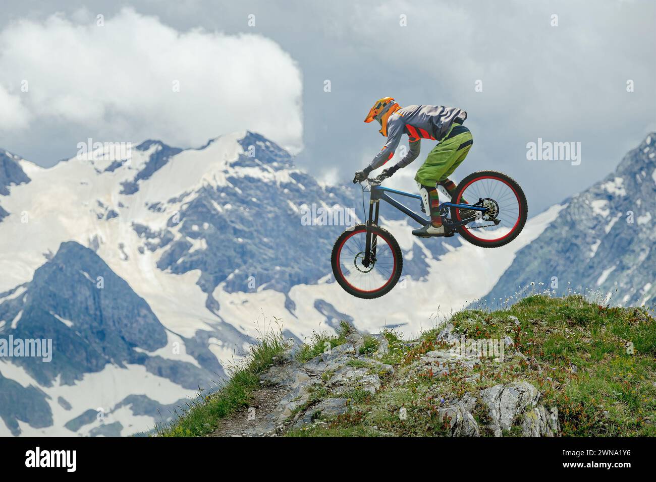 Mountain biker defies gravity, soaring mid-air against backdrop of snow-capped peaks and lush greenery. Ideal for adventure, sports, and nature themes Stock Photo