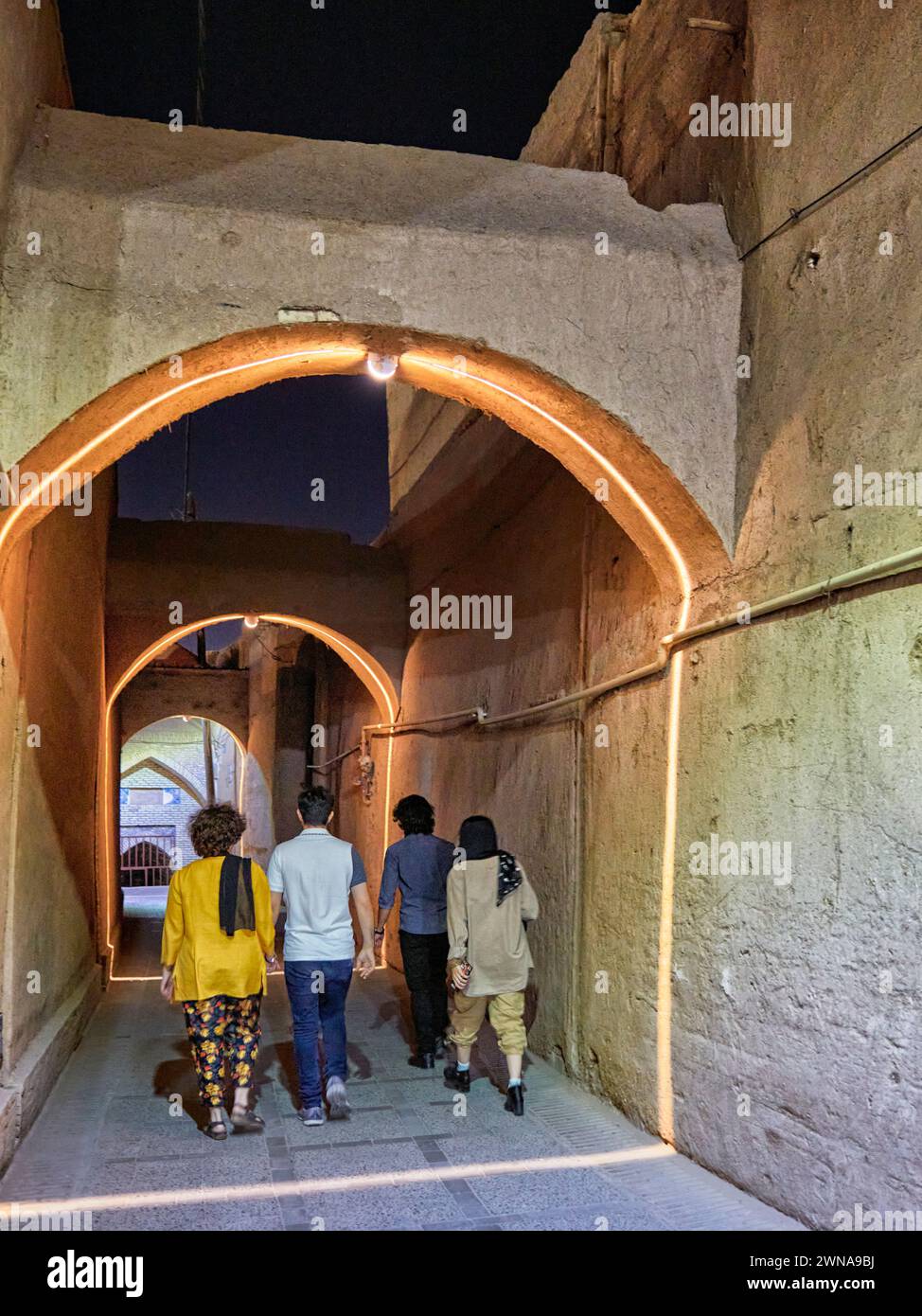 People walk in a narrow street with arches brightly illuminated at night in the historical Fahadan Neighborhood of Yazd, Iran. Stock Photo