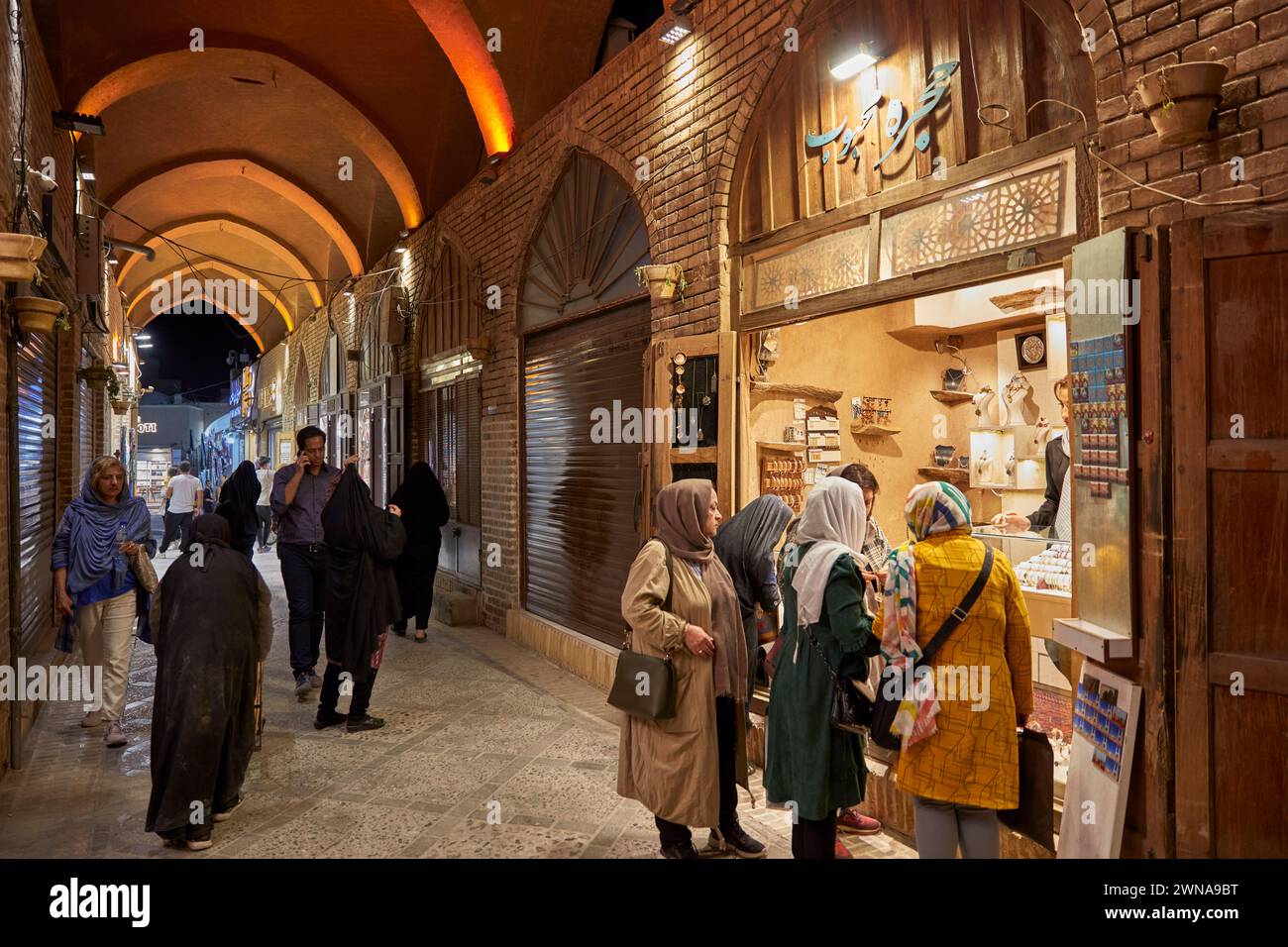 People walk in a busy narrow covered street brightly illuminated at night. Old City of Yazd, Iran. Stock Photo