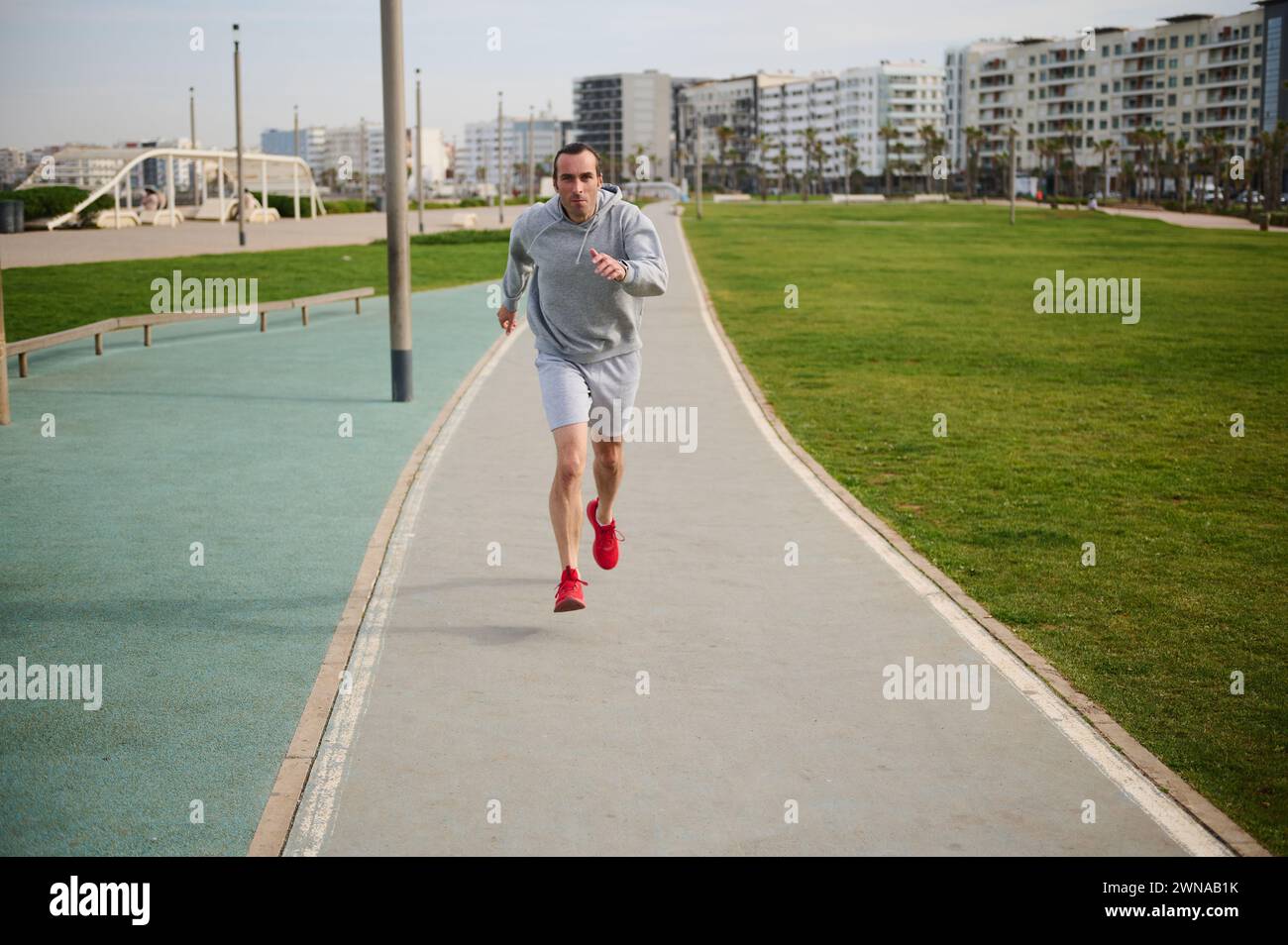 Full length shot of healthy young man in gray sportswear and red sneakers, running on the promenade. Male runner sprinting on the treadmill outdoors. Stock Photo