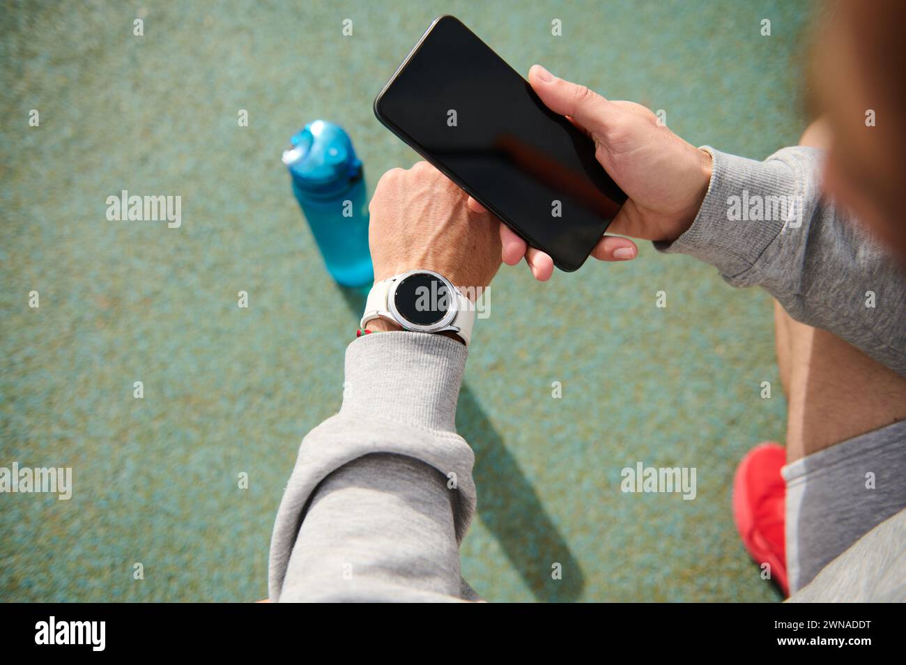 View from above of n athlete holding smart phone with black mockup screen, checking mobile app on his wristwatch, taking pulse or heart rate after hea Stock Photo