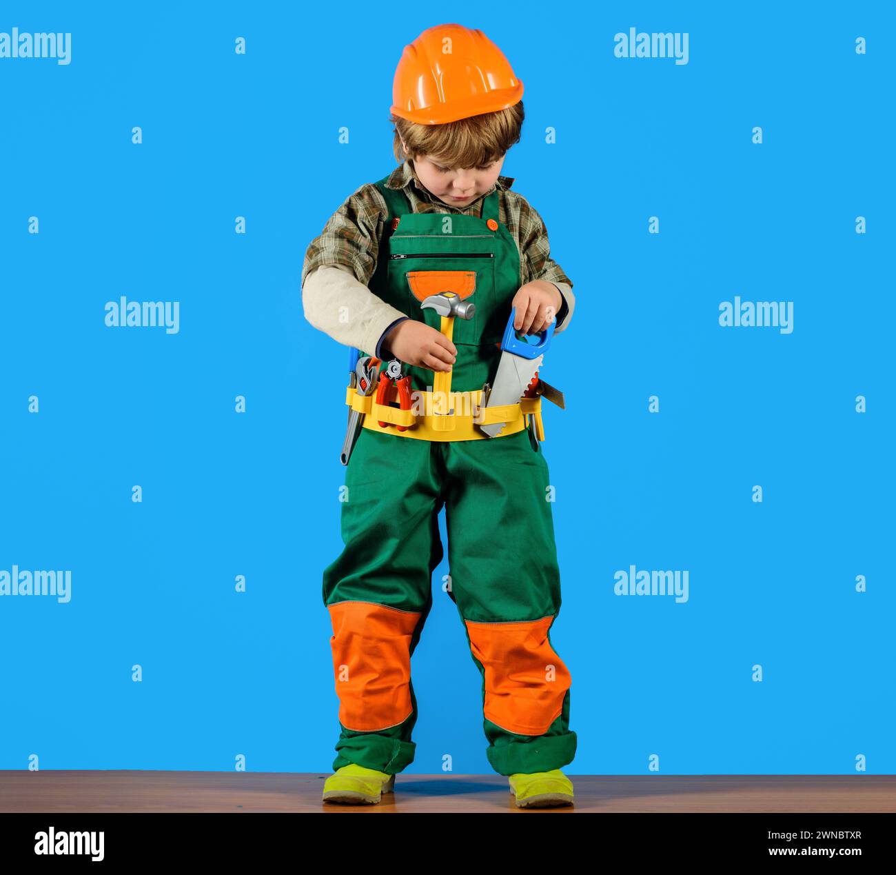 Little builder child playing with toy hammer and saw. Child game. Small boy in overalls and hard hat with builder toy tools set. Cute kid in Stock Photo