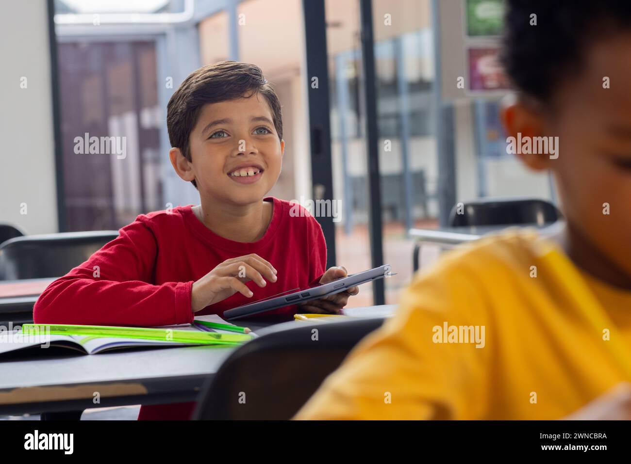 Biracial boy in red smiles with a tablet in a school classroom Stock ...