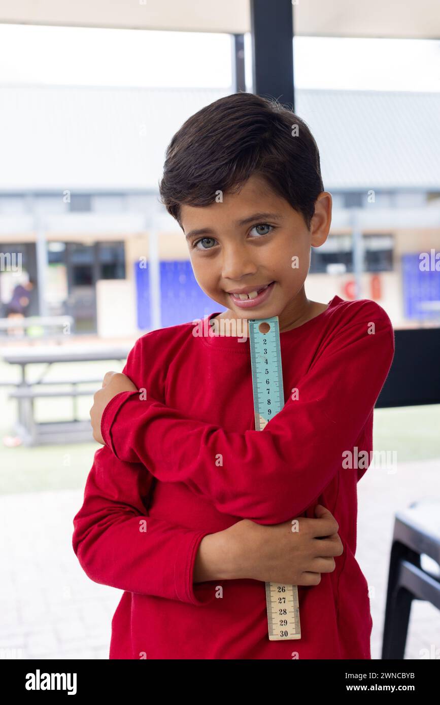 Biracial boy with a bright smile holds a ruler, wearing a red shirt in ...