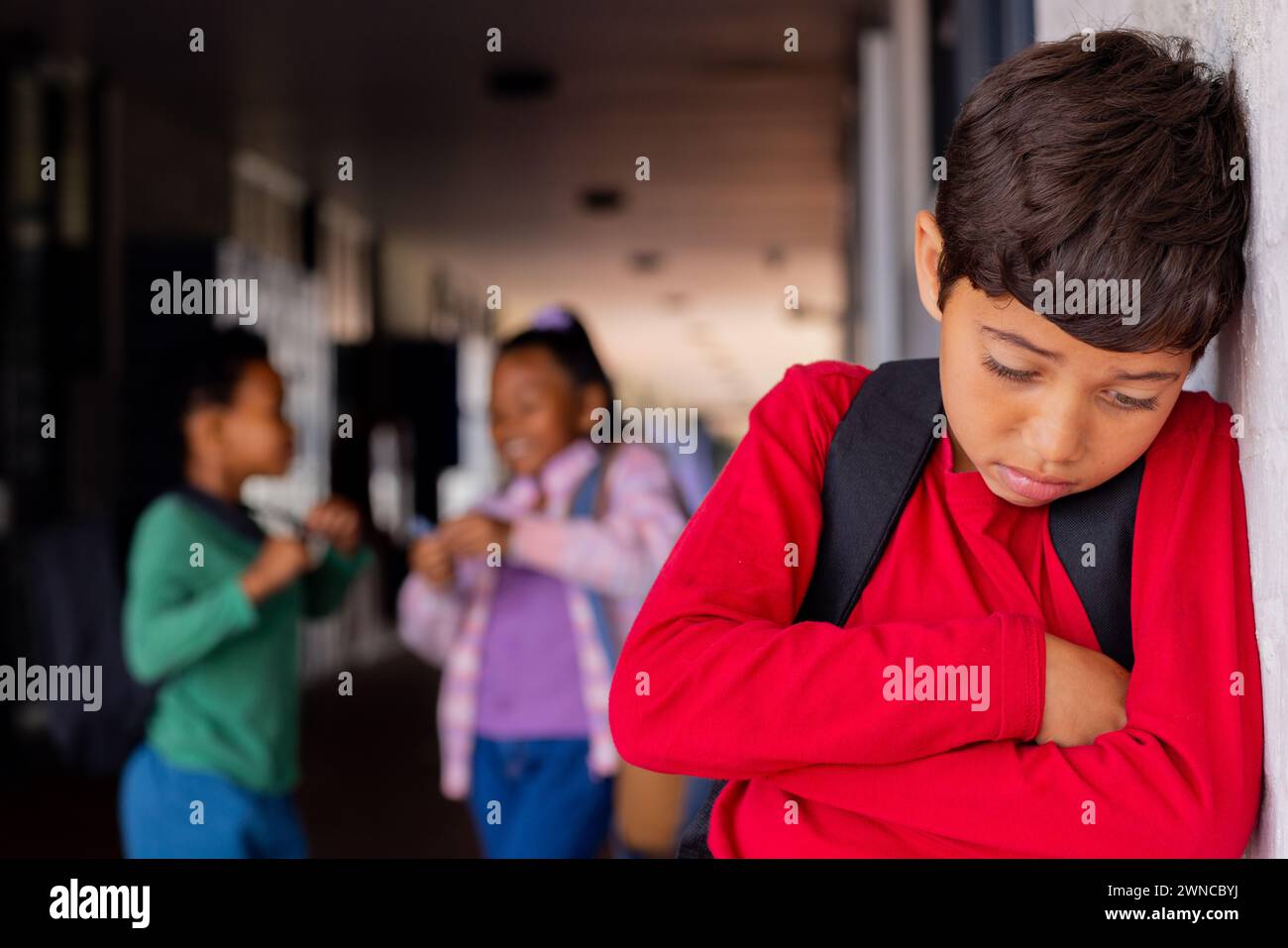 Biracial boy in a red shirt looks upset, leaning against a wall in school, experiencing bullying Stock Photo