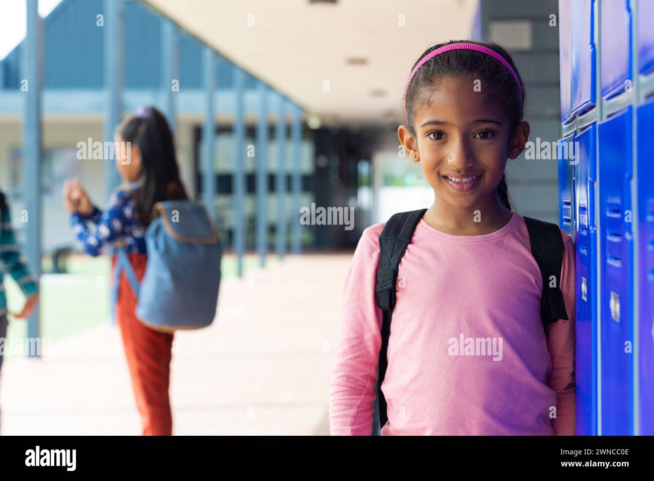 Biracial girl smiles by lockers, another girl in background at school. Stock Photo