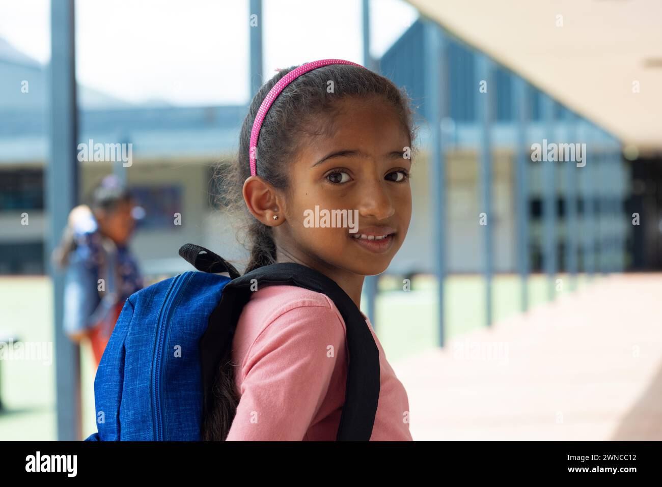 Biracial girl with a pink headband and a blue backpack smiles at school Stock Photo